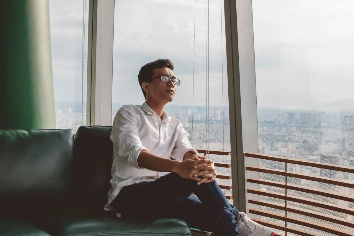 A man sitting by a large window, looking out over the city in quiet reflection.