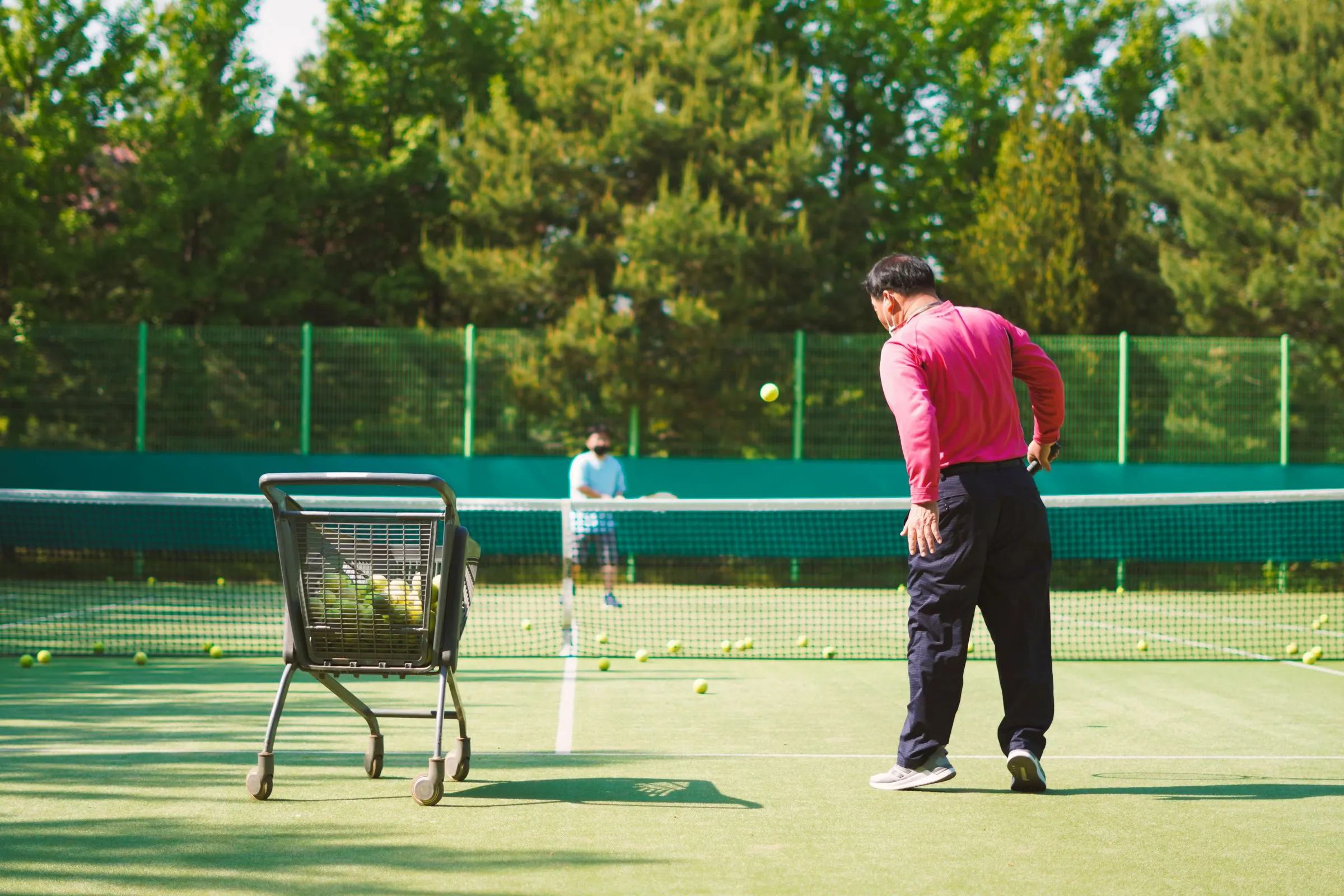 A tennis coach feeding balls from a ball cart to a student across the net during a lesson.