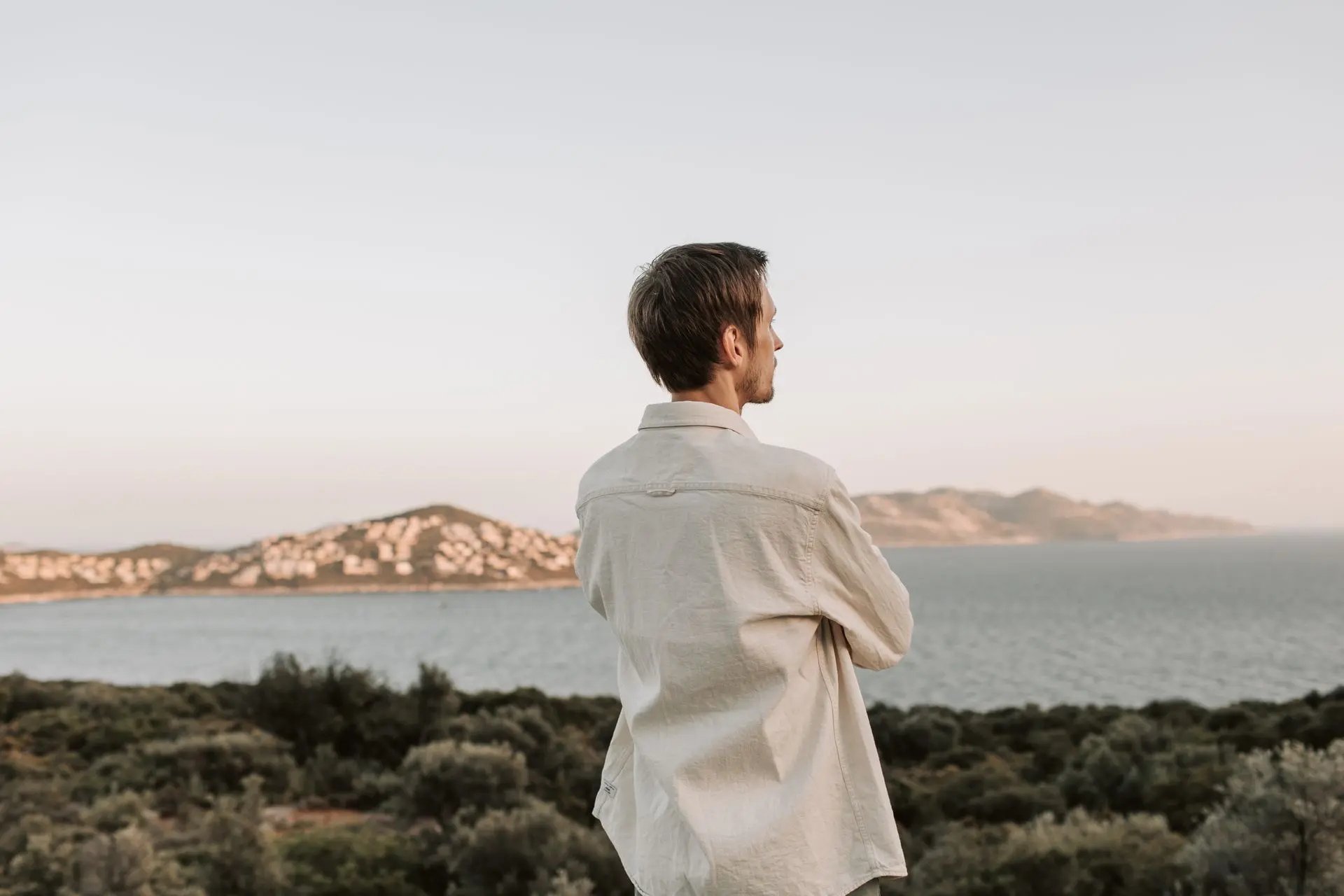A man stands gazing out at the sea and mountains at sunset.